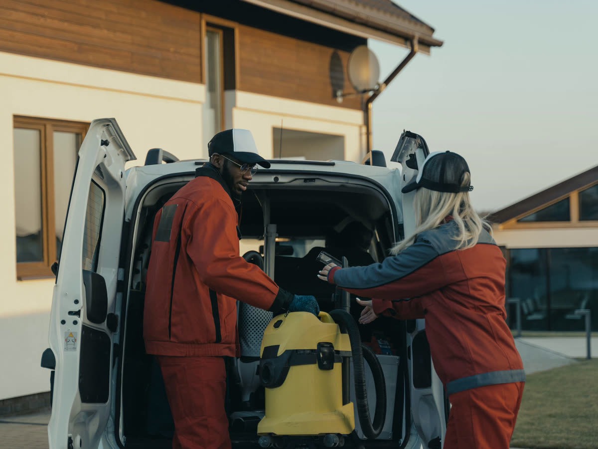 Två städare lastar ur dammsugare och utrustning ur firmabilen utanför ett hus.
